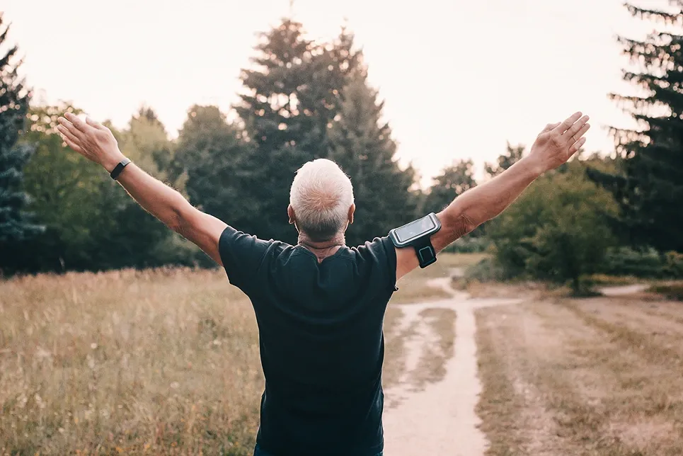 Older man with arms raised outdoors on a sunny trail