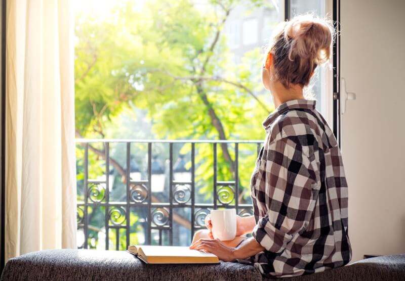 young woman looking through the window while holding a cup of coffee and book